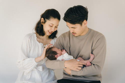 お父さんの腕に抱かれて眠る新生児の赤ちゃんと、隣で微笑むお母さんの家族写真（撮影：モロズミケイタ）
