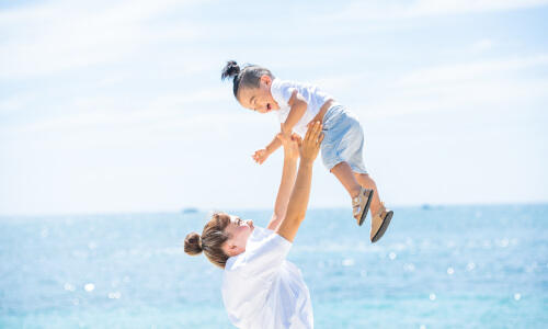 きらきら光る海辺で、高い高いされて楽しそうに笑う1歳の息子さんとそのママの写真(撮影:ワタナベ ナオト)