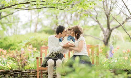 春の花と新緑に囲まれて、ベンチに座ったママとパパと女の子のナチュラルな写真(撮影:SAMIYA)