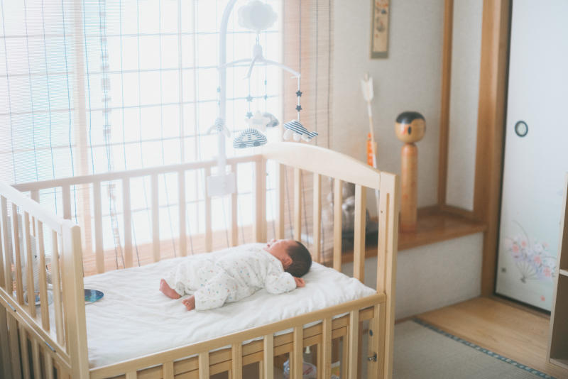  日本の伝統的な和室で、木製のベビーベッドに寝ている新生児。ベッドにはメリーが吊るされ、背景には木製のこけし人形と障子が見える。