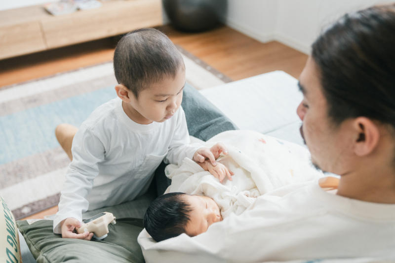 父親に抱かれて眠る白い服を着た赤ちゃんの手を指先で触れる兄の様子