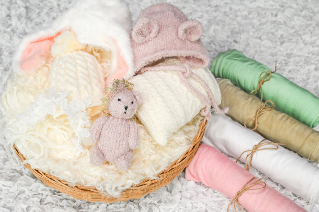 白いおくるみに包まれた赤ちゃん人形の周りをヘッドアクセサリー（黄色とピンクの毛糸の帽子、毛糸のクマのぬいぐるみ、うさぎの耳）と
3色のおくるみで囲んだニューボンフォト