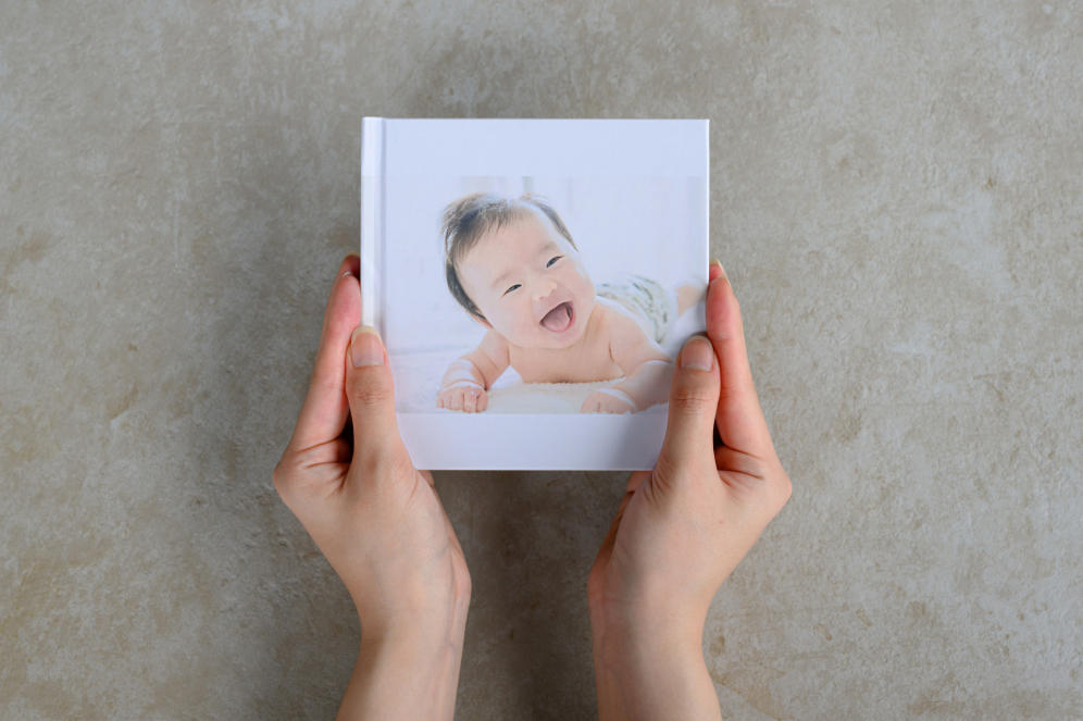 うつ伏せで顔を上げて笑う乳児の写真が表紙になったフォトブックを両手で持つ手元の写真